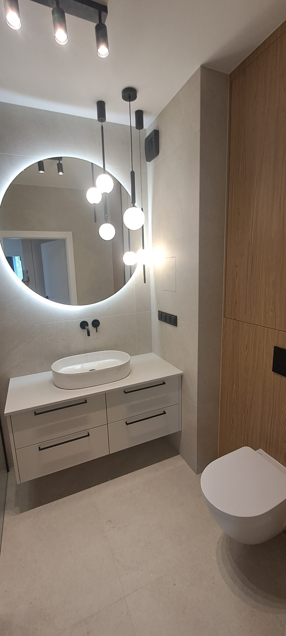 Nowoczesna łazienka z okrągłym lustrem LED, wiszącą szafką z umywalką i toaletą. Jasne płytki i drewniane akcenty tworzą minimalistyczny wystrój. Oświetlenie punktowe.