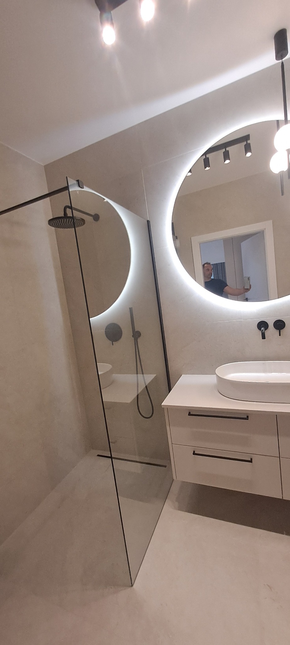 Nowoczesna łazienka z kabiną prysznicową typu walk-in, okrągłym lustrem z podświetleniem LED i białą szafką z umywalką nablatową. Widoczne czarne baterie.