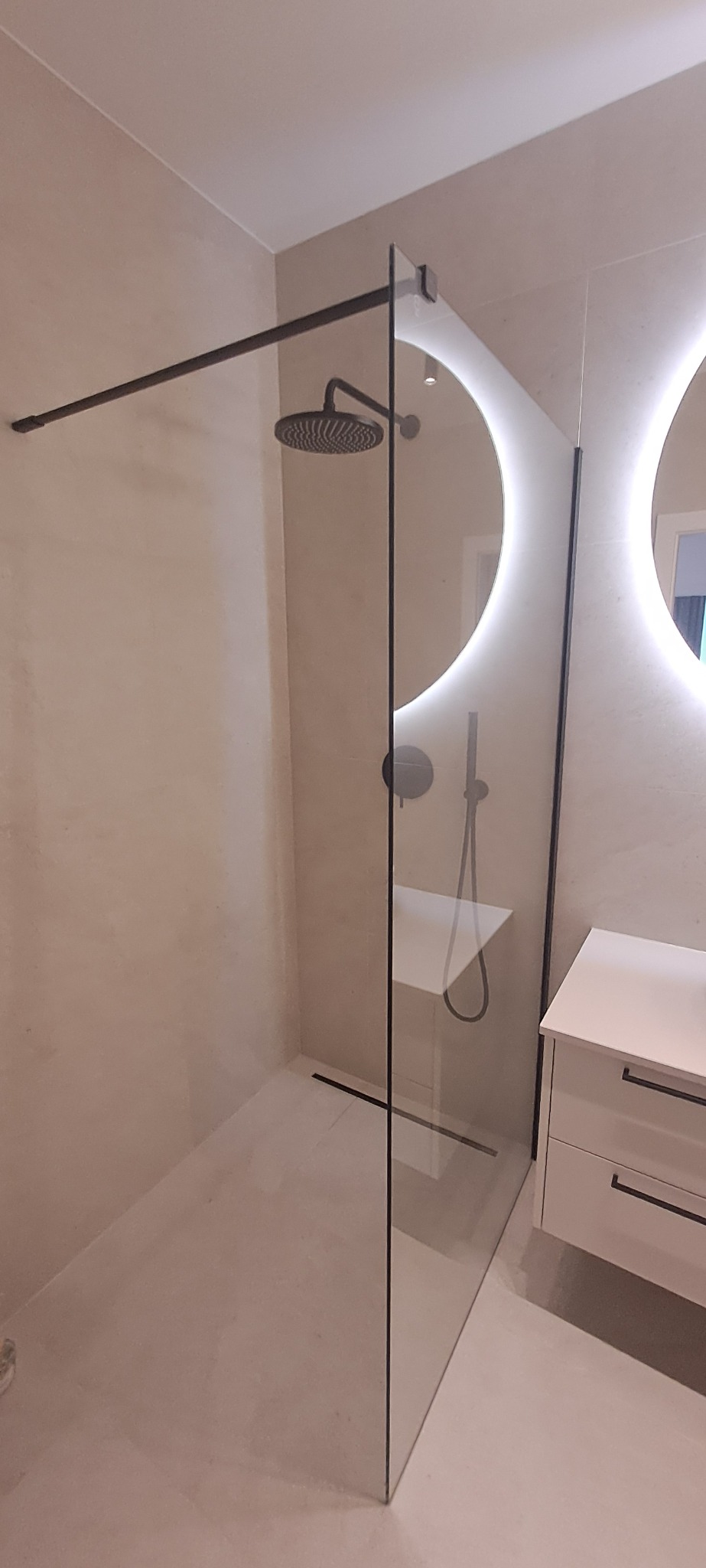 Nowoczesna kabina prysznicowa walk-in z czarnymi elementami, minimalistyczna armatura, oświetlenie LED w lustrach. Jasne płytki i biała szafka umywalkowa.