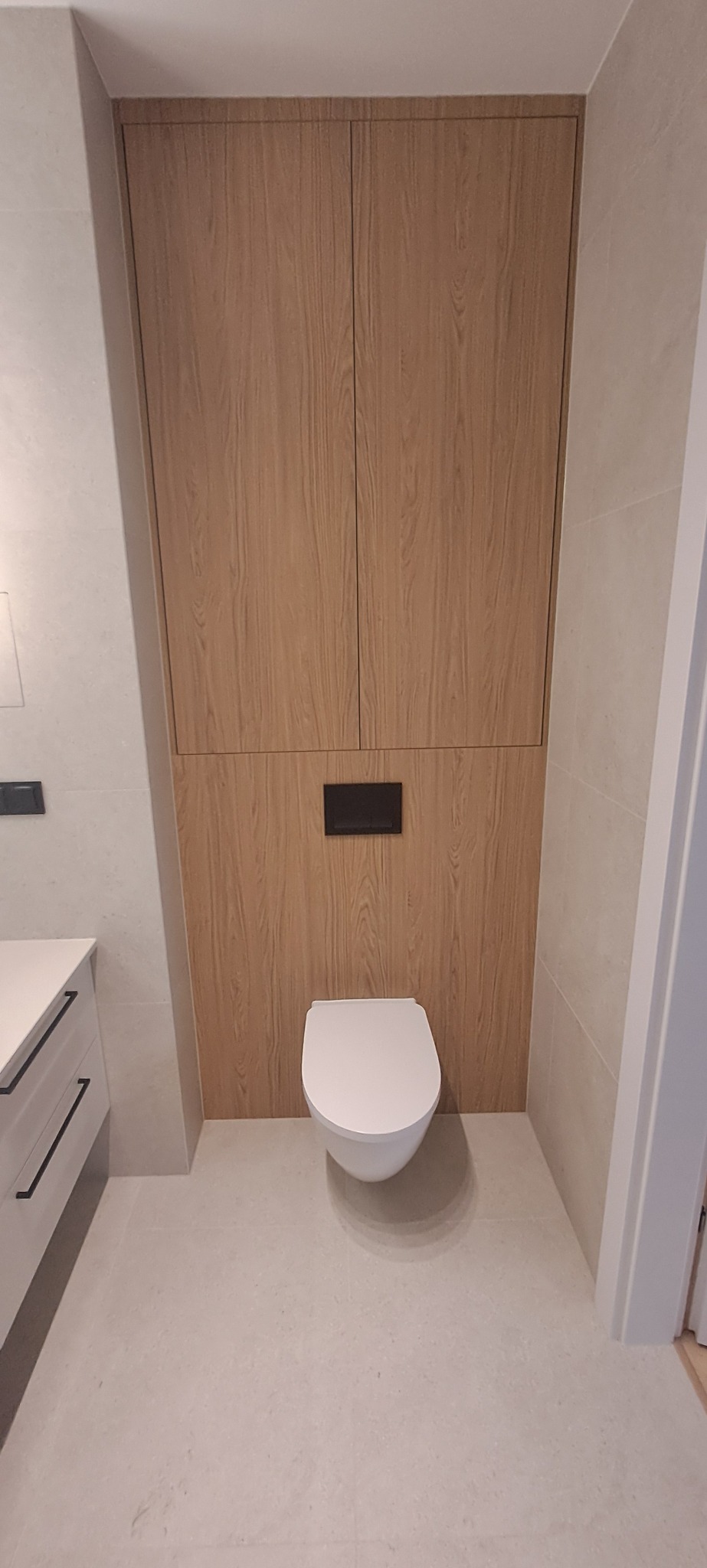 Nowoczesna łazienka z podwieszaną toaletą i zabudową z drewna. Minimalistyczny design, jasne płytki i czarne akcenty. Górna szafka zamykana.