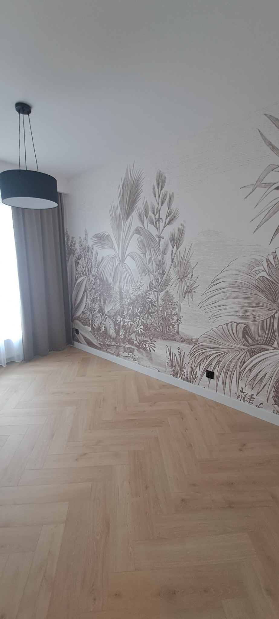 Jasny parkiet ułożony we wzór jodełki francuskiej. Na ścianie duży, dekoracyjny mural z motywem roślin tropikalnych. Minimalistyczna lampa wisząca.