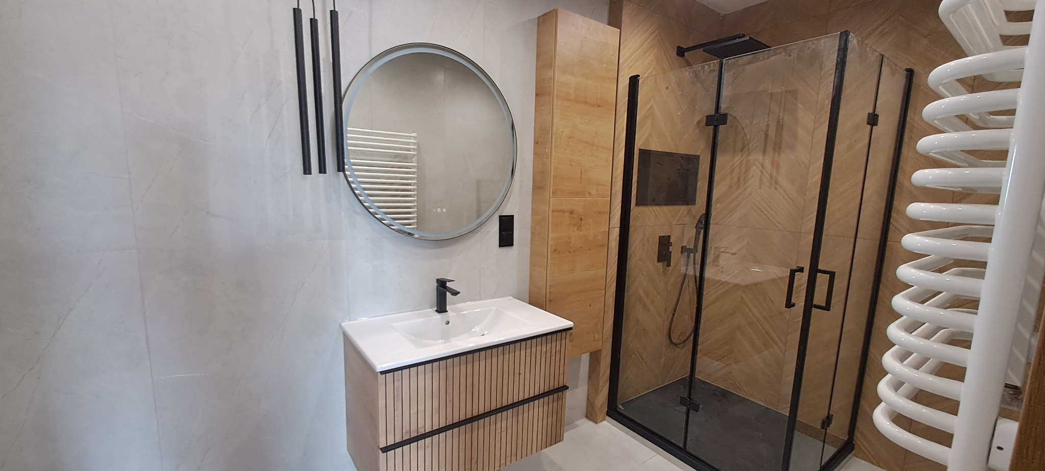 Nowoczesna łazienka po remoncie: umywalka z szafką z lamelami, okrągłe lustro z podświetleniem, kabina prysznicowa z czarnymi ramami i grzejnik.