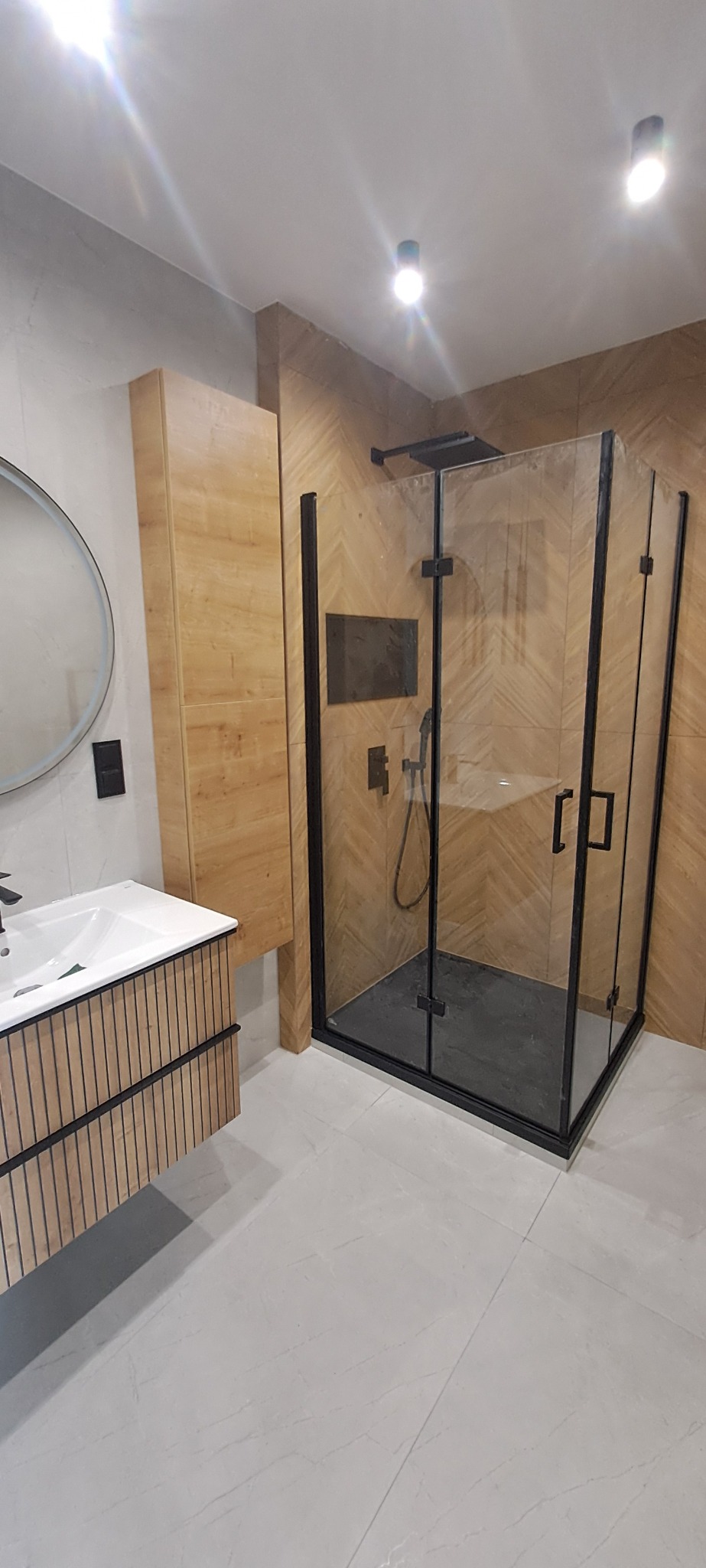 Nowoczesna łazienka z kabiną prysznicową typu walk-in z czarnymi profilami, drewnianą szafką pod umywalką i płytkami imitującymi drewno na ścianie.