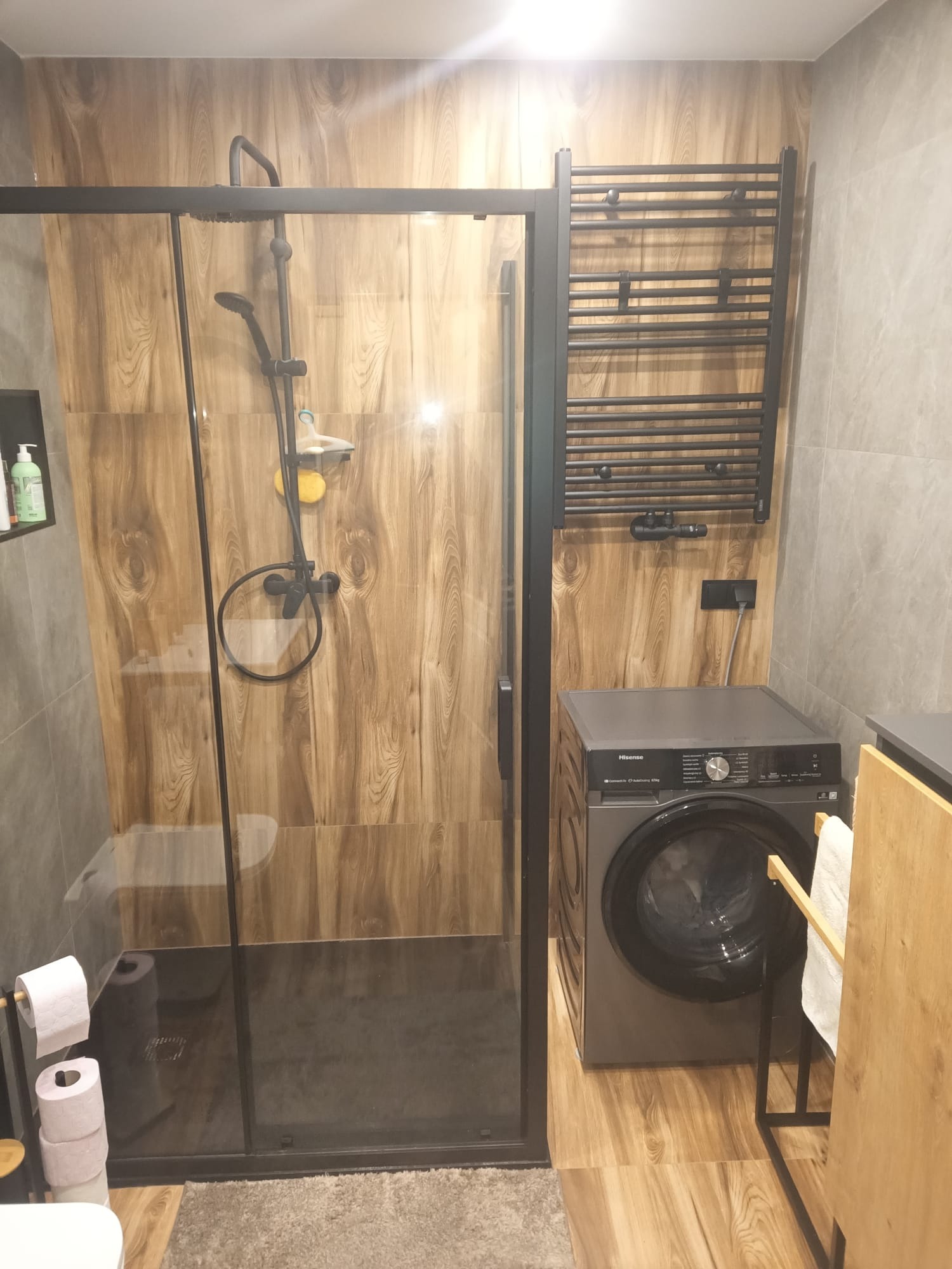 Nowoczesna łazienka z kabiną prysznicową typu walk-in, grzejnikiem drabinkowym i pralką. Ściany wyłożone płytkami imitującymi drewno i szarym kamieniem.