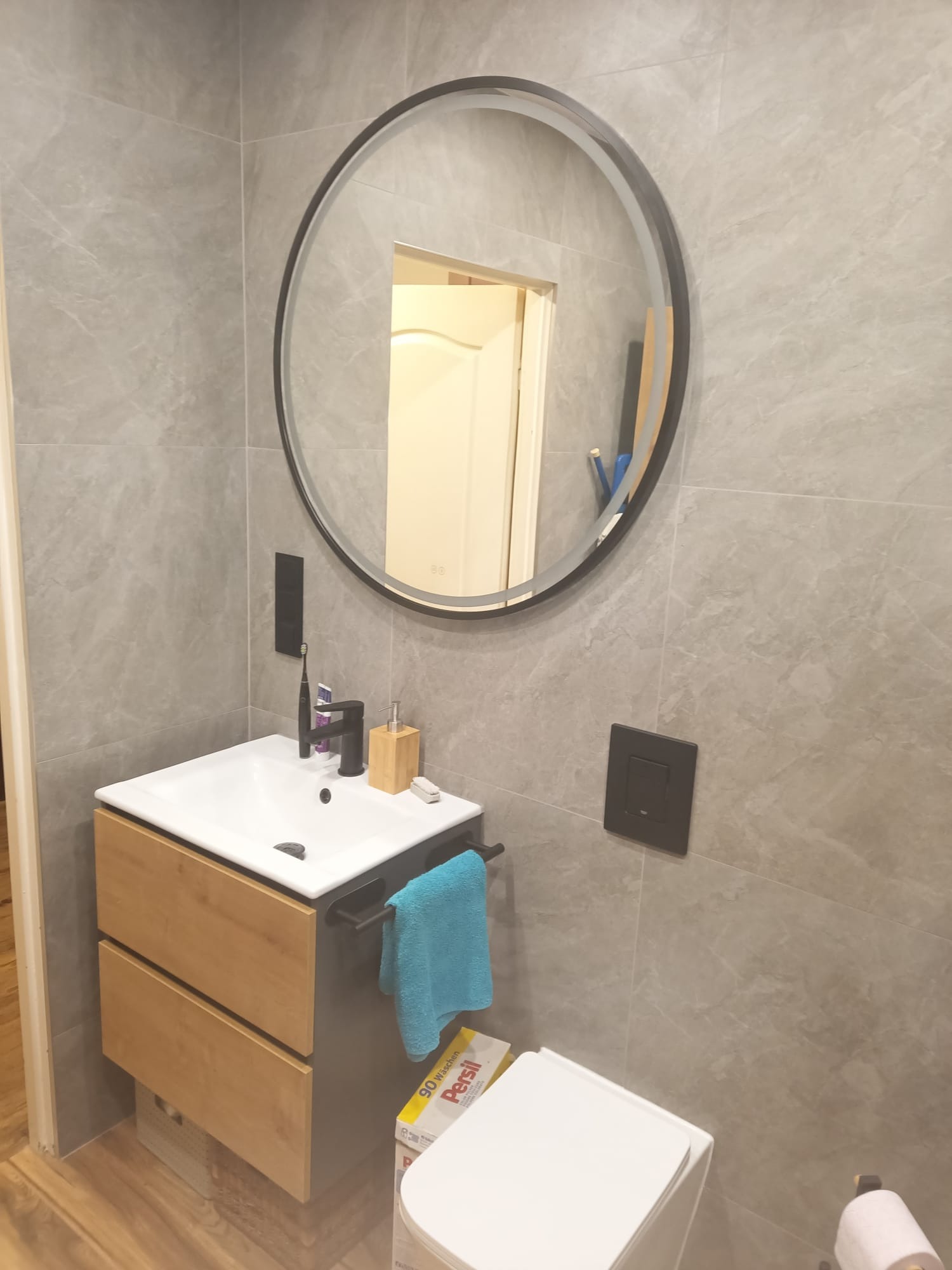 Nowoczesna łazienka z okrągłym lustrem z podświetleniem LED, szarą płytką imitującą kamień i drewnianą szafką pod umywalką. Widoczny fragment toalety i akcesoria łazienkowe.