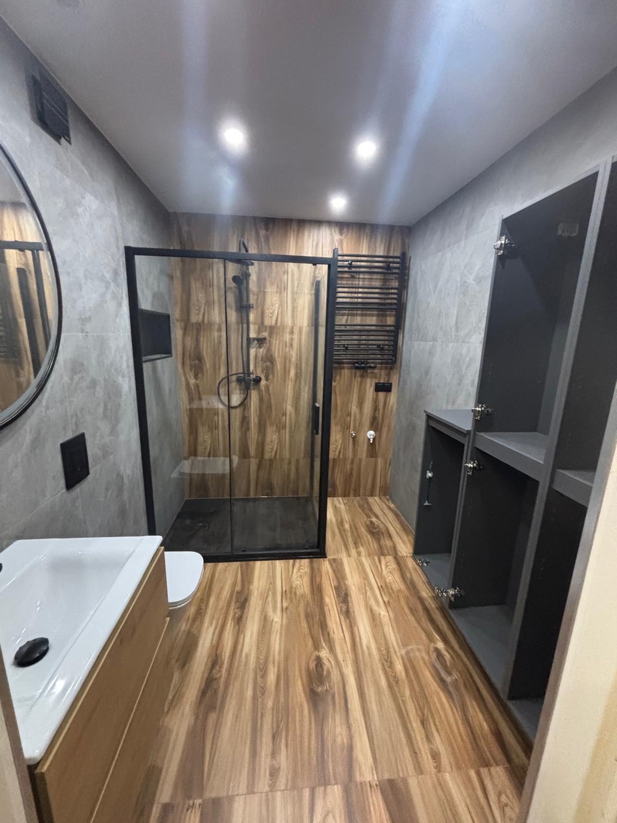 Nowoczesna łazienka z kabiną prysznicową typu walk-in, drewnopodobną podłogą i szarą armaturą. Widoczna umywalka z szafką i grzejnik drabinkowy.