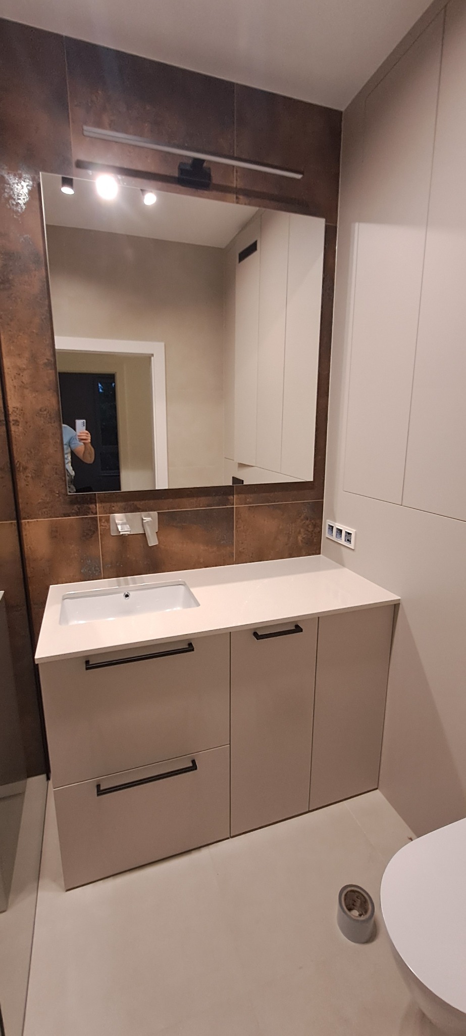 Nowoczesna łazienka z szafką pod umywalką w odcieniach beżu i brązu. Uwagę przykuwają ciemne uchwyty i lustro z oświetleniem LED. Widoczny fragment toalety.