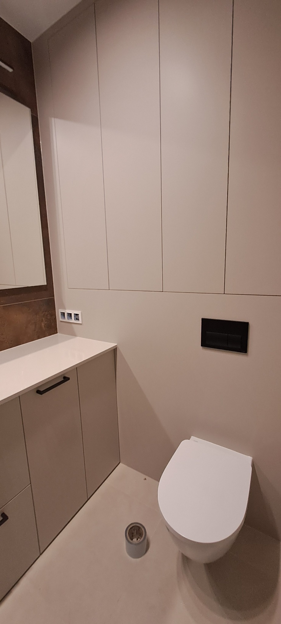 Nowoczesna łazienka po remoncie: podwieszana toaleta, zabudowa meblowa w jasnym odcieniu, minimalistyczny design, czarny przycisk spłukiwania, lustro z brązową ramą.