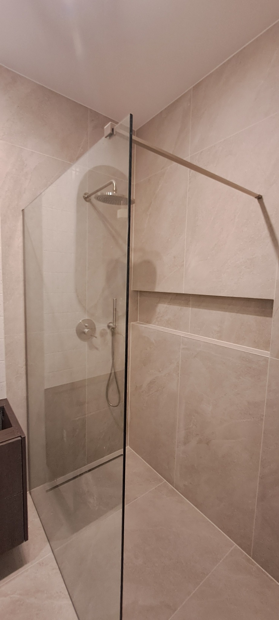 Nowoczesna kabina prysznicowa walk-in z deszczownicą i szklaną ścianką, wykończona beżowymi płytkami imitującymi kamień, z wnęką w ścianie. Minimalistyczny design.