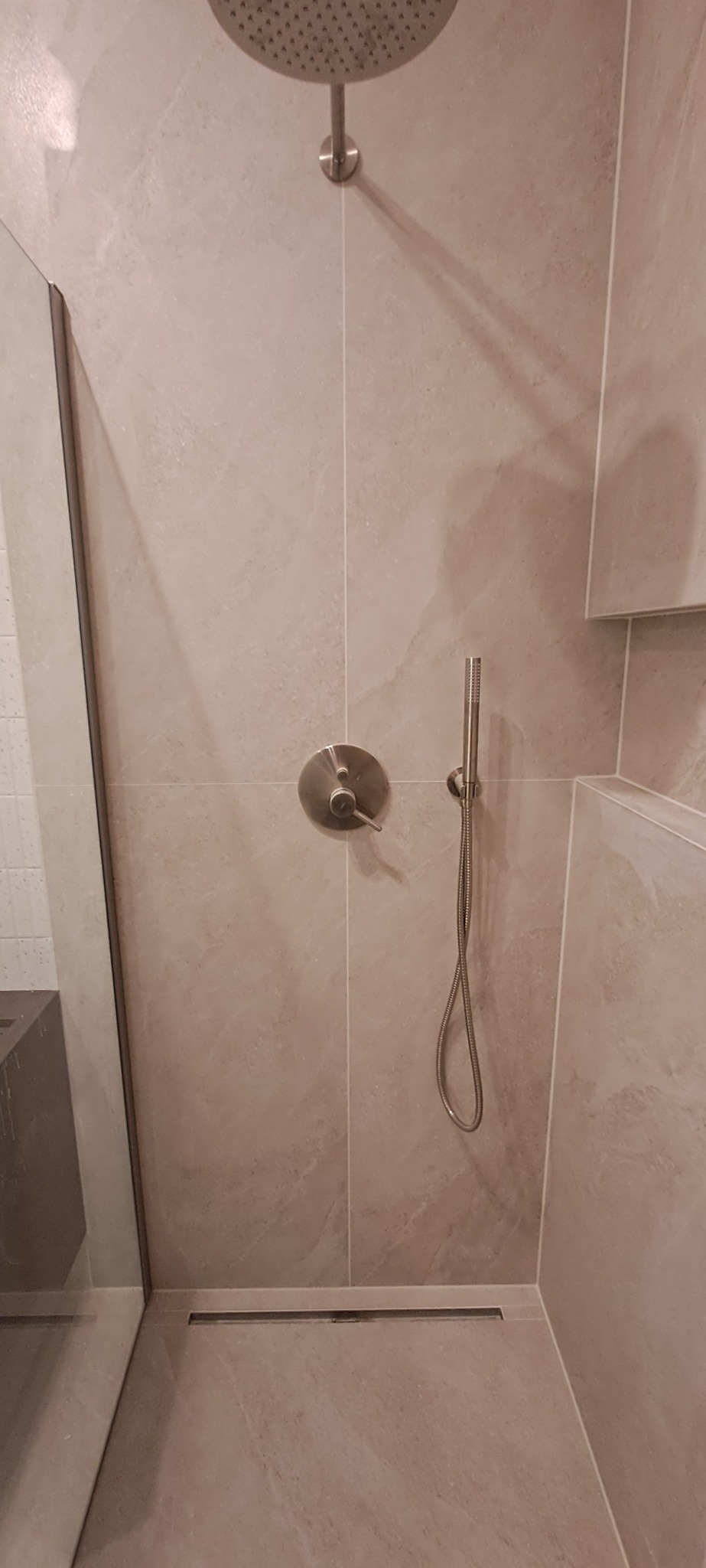 Beżowa kabina prysznicowa z deszczownicą i odpływem liniowym. Minimalistyczny design, płytki imitujące kamień, szklana ściana. Nowoczesne wykończenie łazienki.