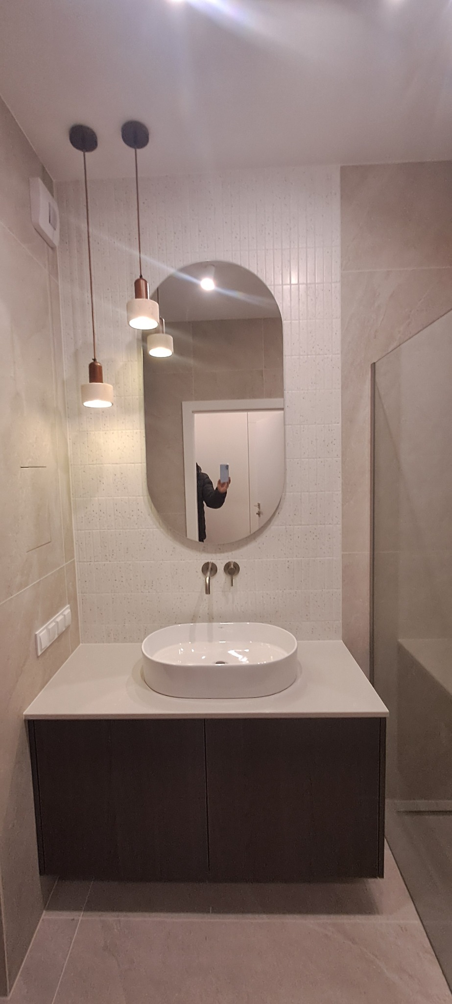 Nowoczesna łazienka z owalnym lustrem, wiszącą szafką z umywalką i designerskimi lampami. Jasne płytki na ścianach i podłodze. Minimalistyczny styl.