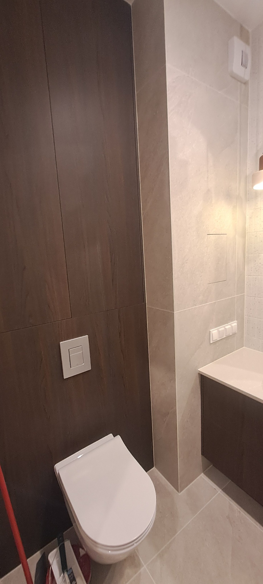 Nowoczesna łazienka po remoncie: podwieszana toaleta z brązową ścianą, beżowe płytki, minimalistyczna umywalka z szafką. Widoczny fragment mopa.