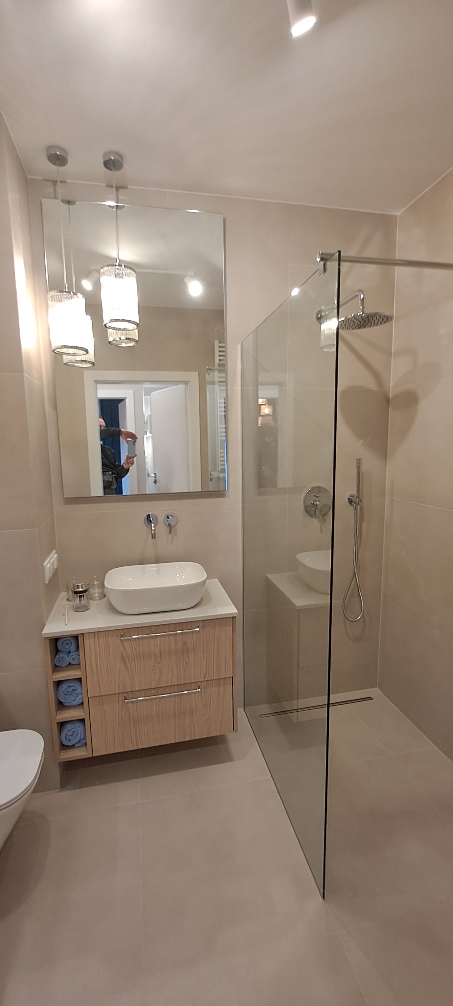 Jasna łazienka z kabiną prysznicową typu walk-in, umywalką nablatową na szafce z drewnianymi frontami i lustrem z ozdobnymi lampami wiszącymi.