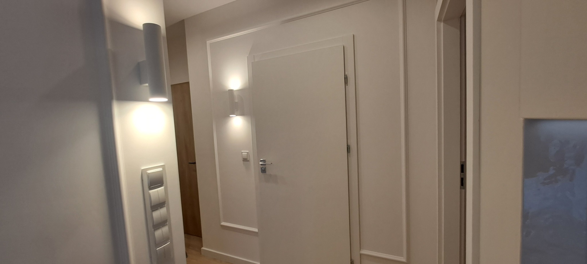 Białe drzwi wewnętrzne z ozdobnymi listwami, oświetlenie punktowe na ścianie, widok na fragment korytarza z drewnianymi drzwiami w tle. Minimalistyczny design.