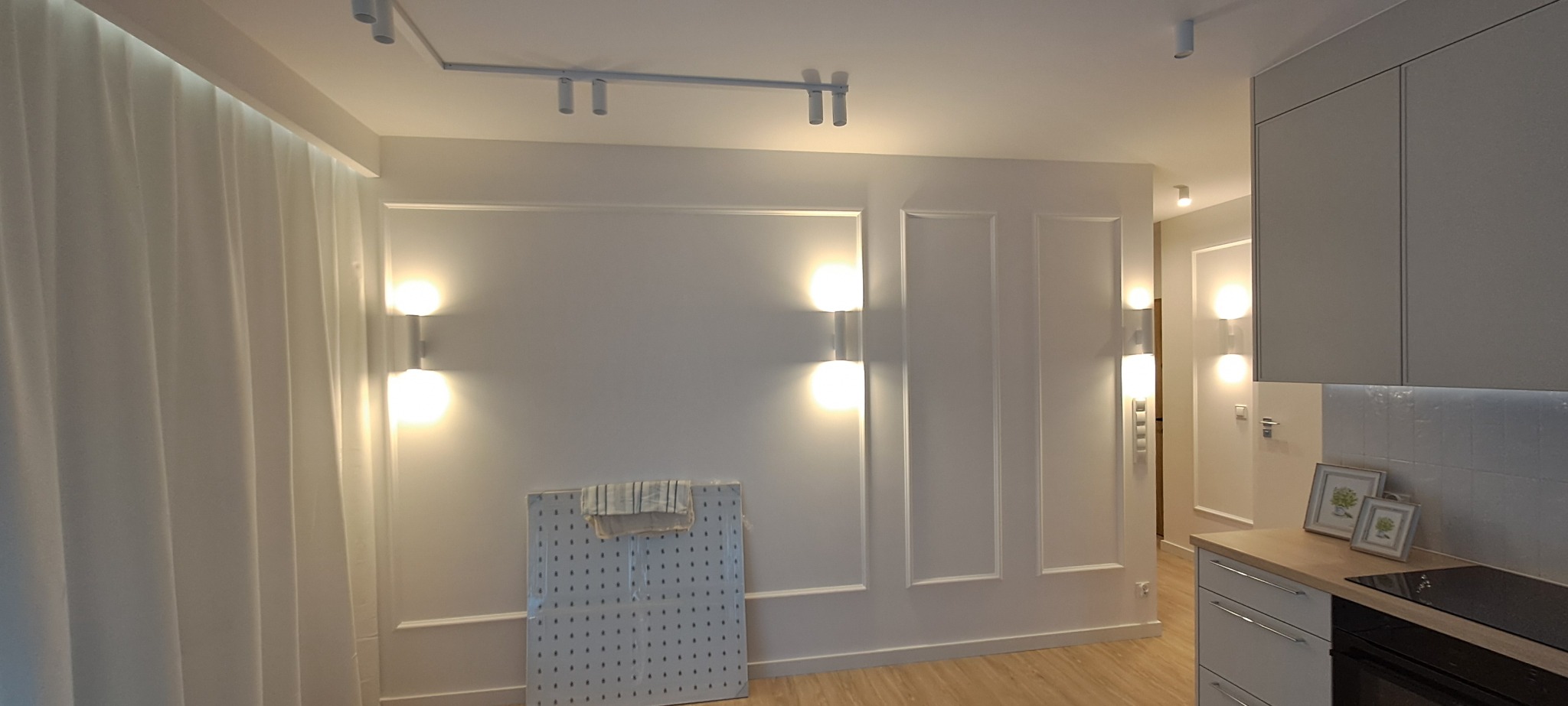 Jasne wnętrze po remoncie: białe ściany z dekoracyjnymi panelami, nowoczesne oświetlenie, minimalistyczna kuchnia z drewnianym blatem i subtelne dekoracje.