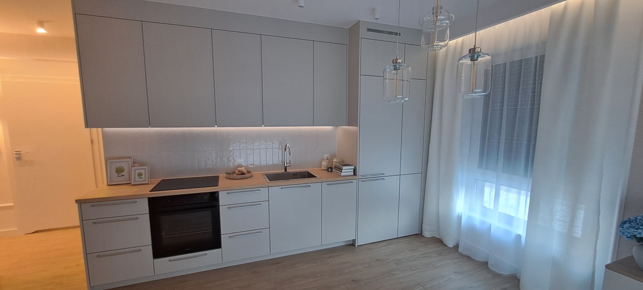 Jasna kuchnia z szarymi szafkami, drewnianym blatem i trzema szklanymi lampami wiszącymi. Minimalistyczny design z białymi zasłonami i drewnianą podłogą.