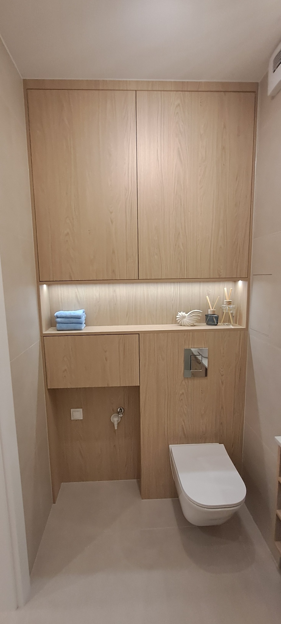 Nowoczesna łazienka z zabudową meblową z drewna, podświetlana półka z dekoracjami i toaleta podwieszana. Minimalistyczny design, ciepłe oświetlenie.