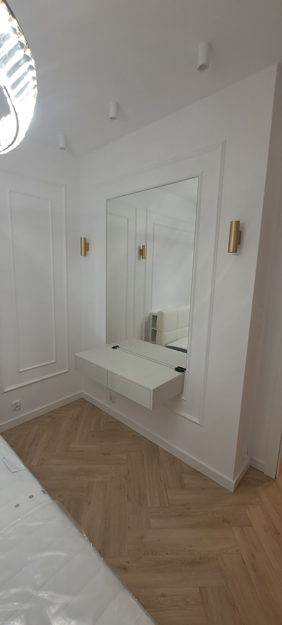 Elegancka toaletka z lustrem w białej ramie, zamontowana na ścianie w pokoju z parkietem ułożonym w jodełkę. Dwa złote kinkiety po bokach lustra.