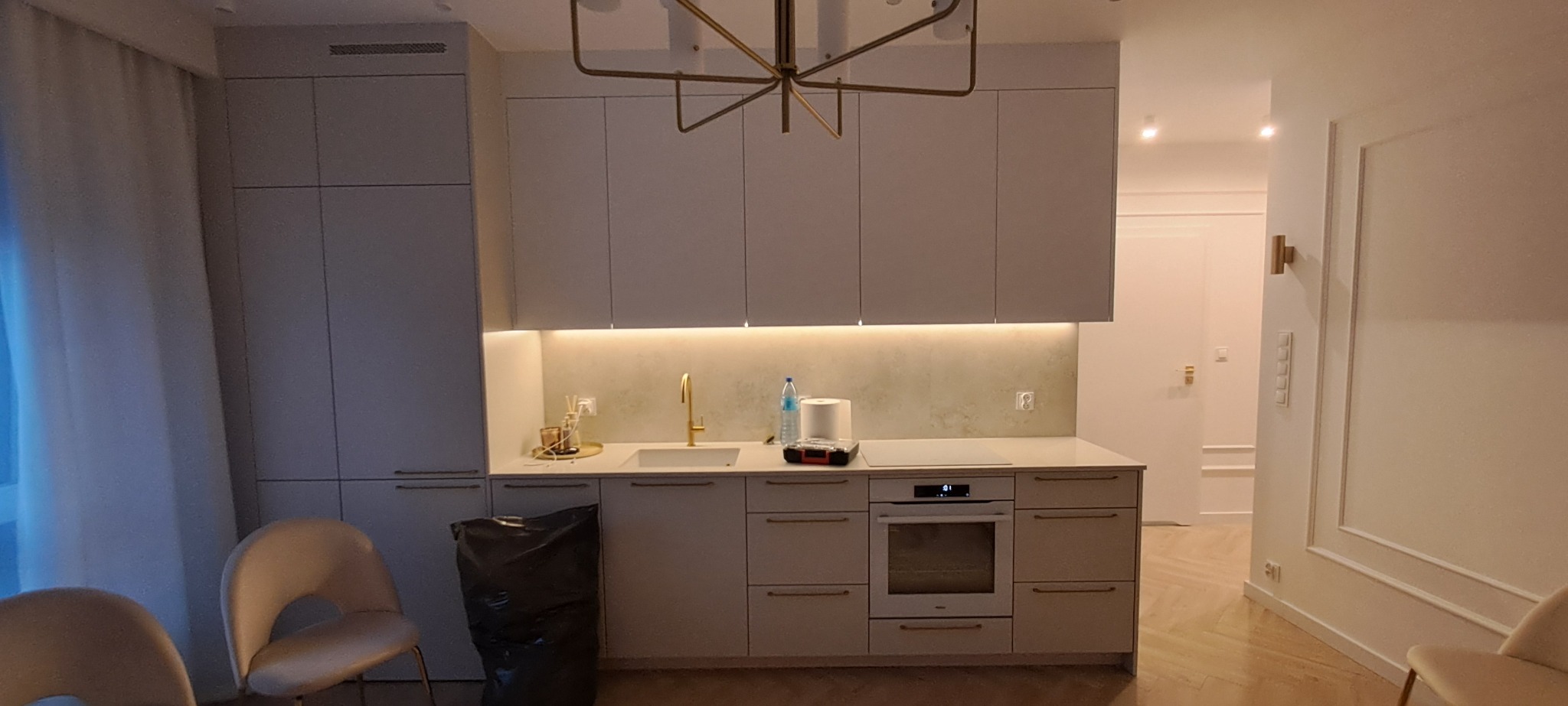 Jasna kuchnia z oświetleniem LED podszafkowym i złotymi akcentami, widok na sąsiednie pomieszczenie z dekoracyjną sztukaterią. Uwagę zwraca nowoczesny żyrandol.