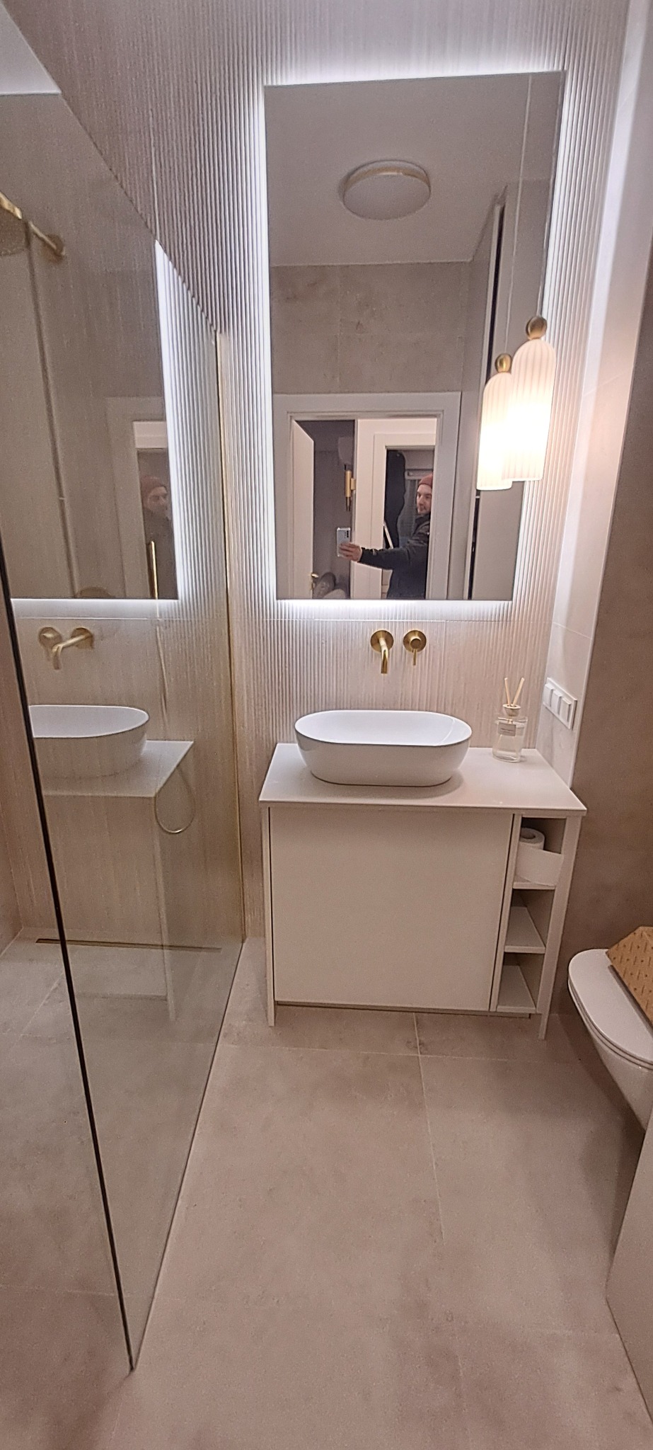 Nowoczesna łazienka z umywalką nablatową, lustrem z podświetleniem LED i złotą armaturą. Widoczne płytki z delikatnym wzorem i minimalistyczne oświetlenie.