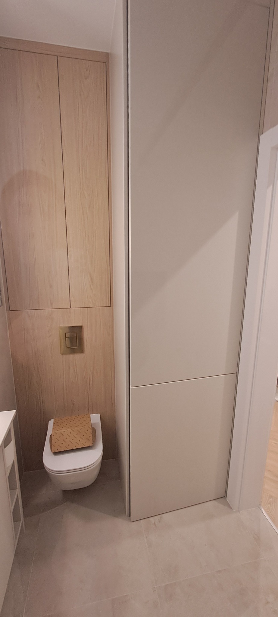 Nowoczesna łazienka z zabudową WC w odcieniach beżu i drewna. Minimalistyczny design, wisząca miska WC i ukryta spłuczka. Estetyczne wykończenie wnętrza.