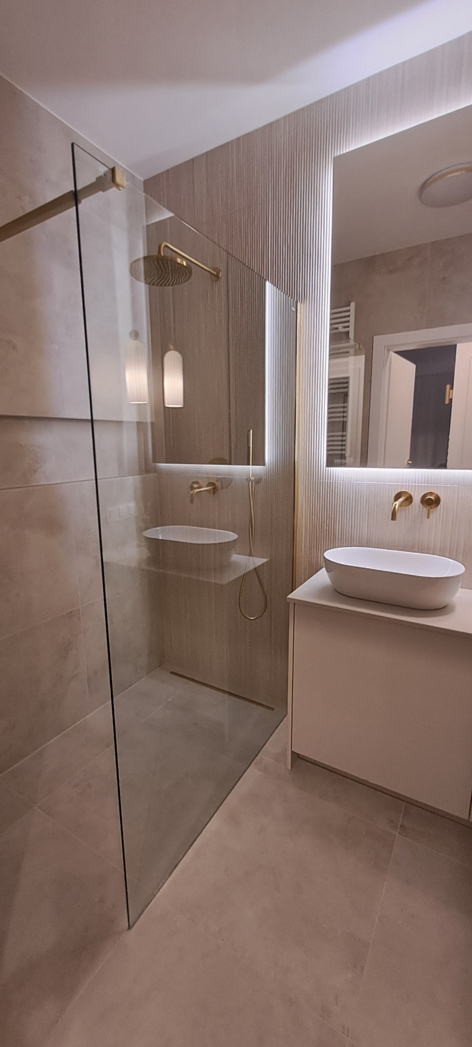 Nowoczesna łazienka z kabiną typu walk-in, złotą armaturą i umywalką nablatową. Ściany wykończone płytkami w odcieniach beżu z subtelnym, pionowym wzorem.