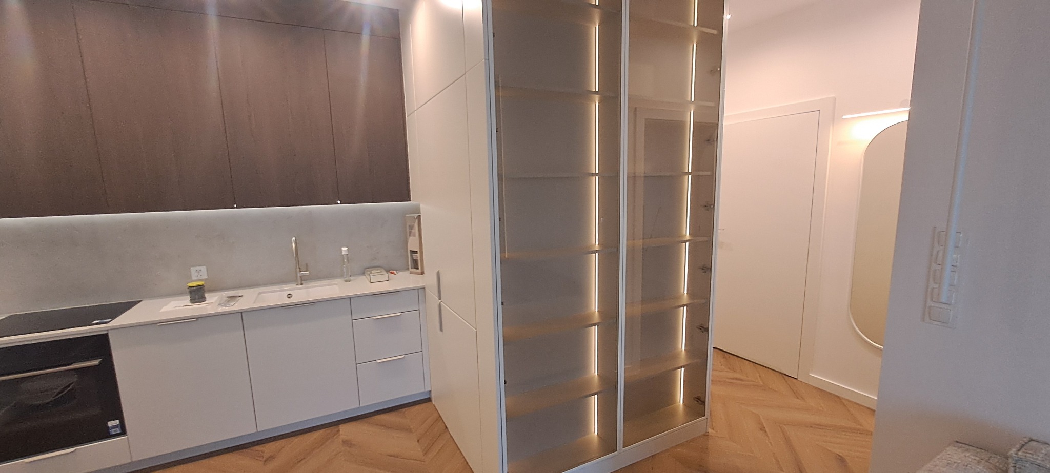 Nowoczesna kuchnia z zabudową meblową w odcieniach bieli i szarości, z podświetlanymi półkami i drewnianą podłogą w jodełkę. Minimalistyczny design, jasne wnętrze.