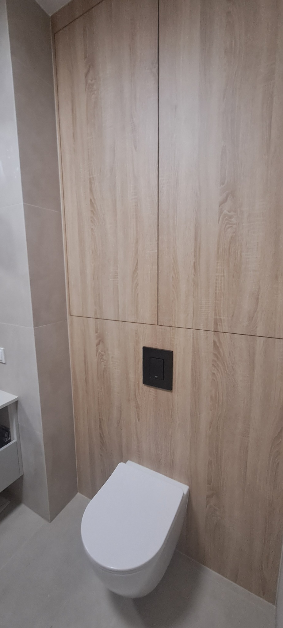 Nowoczesna toaleta podwieszana z zabudową meblową w odcieniach drewna. Czarny przycisk spłuczki na tle drewnianej ściany. Minimalistyczny design łazienki.