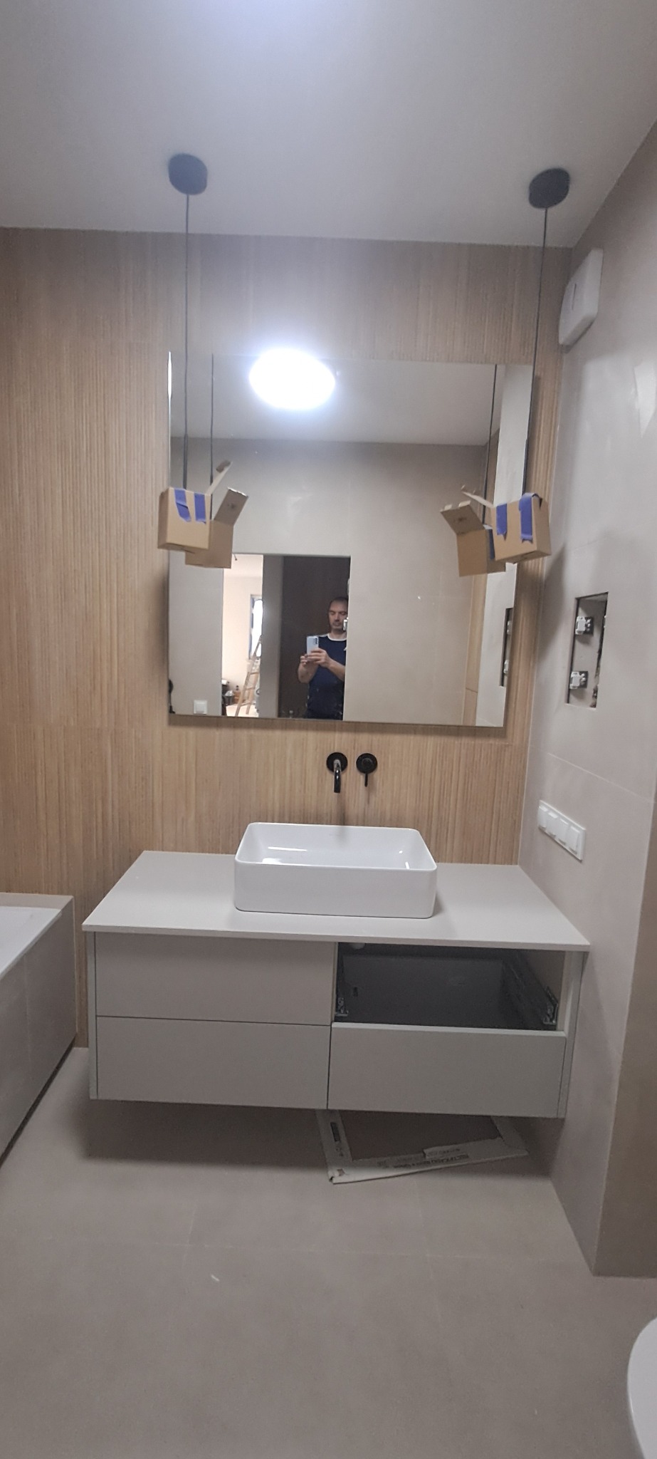 Nowoczesna łazienka z umywalką nablatową i szafką w odcieniach bieli i beżu. W lustrze odbija się osoba robiąca zdjęcie. Widoczne kartony zabezpieczające lampy.