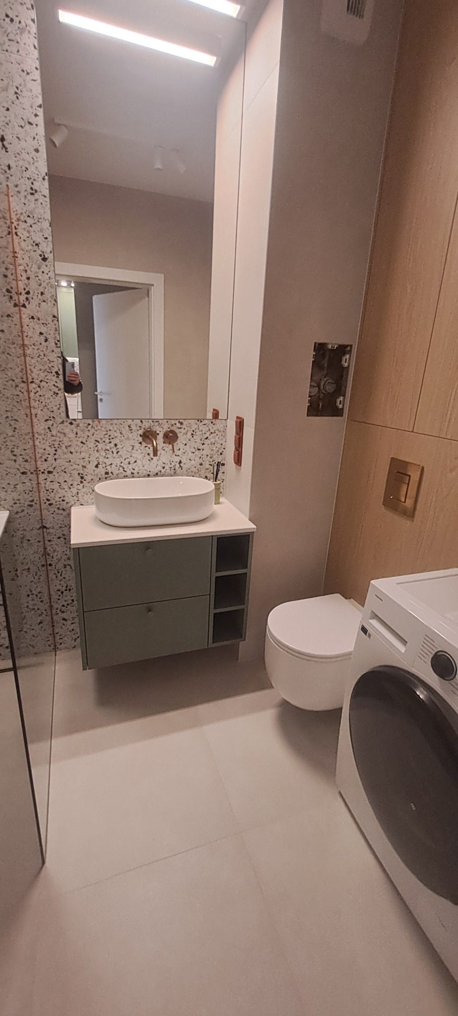 Nowoczesna łazienka z umywalką nablatową, szafką w kolorze oliwkowym, toaletą i pralką. Ściana z mozaiką i lustro z oświetleniem LED. Minimalistyczny design.