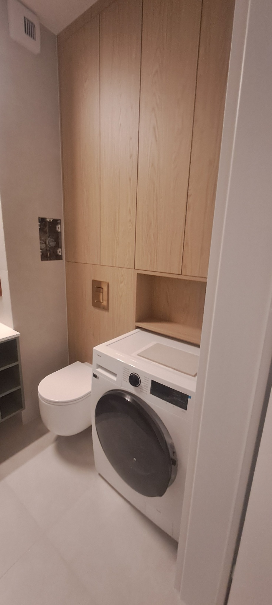 Jasna łazienka z drewnianą zabudową meblową, toaletą podwieszaną i pralką z umywalką na blacie. Minimalistyczny design, neutralna kolorystyka.