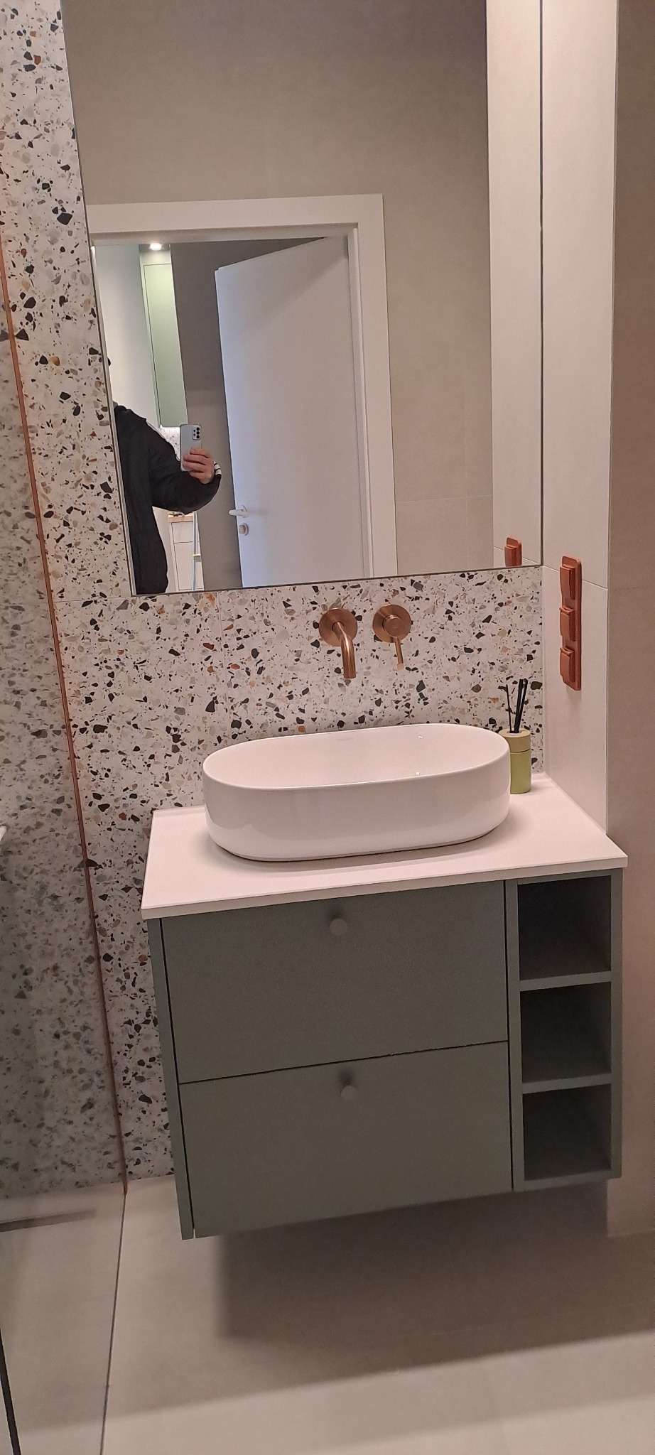 Nowoczesna łazienka z umywalką nablatową, baterią w kolorze miedzi i szafką w odcieniach szarości. Na ścianie mozaika z drobnymi kamyczkami. W lustrze odbija się osoba robiąca zdjęcie.