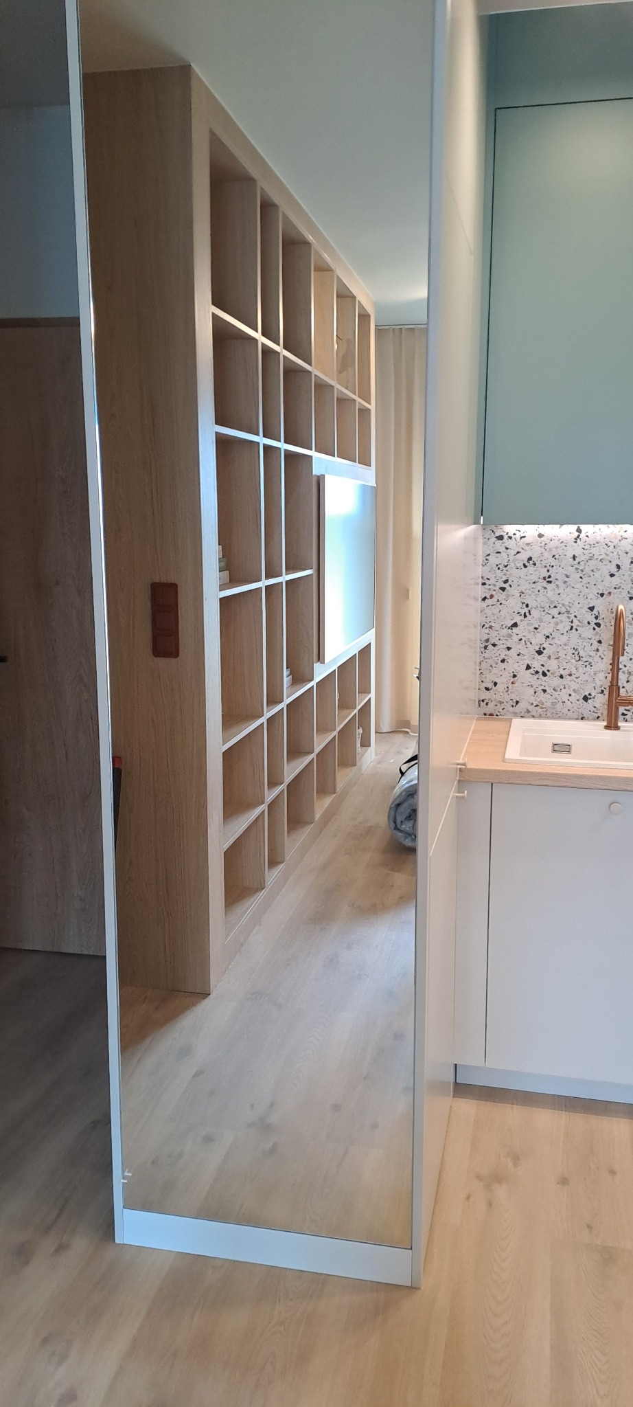 Nowoczesne wnętrze z zabudową kuchenną w bieli i drewnianym regałem z półkami, odbijające się w lustrzanej ścianie. Uwagę zwraca mozaika nad blatem i minimalistyczna armatura.