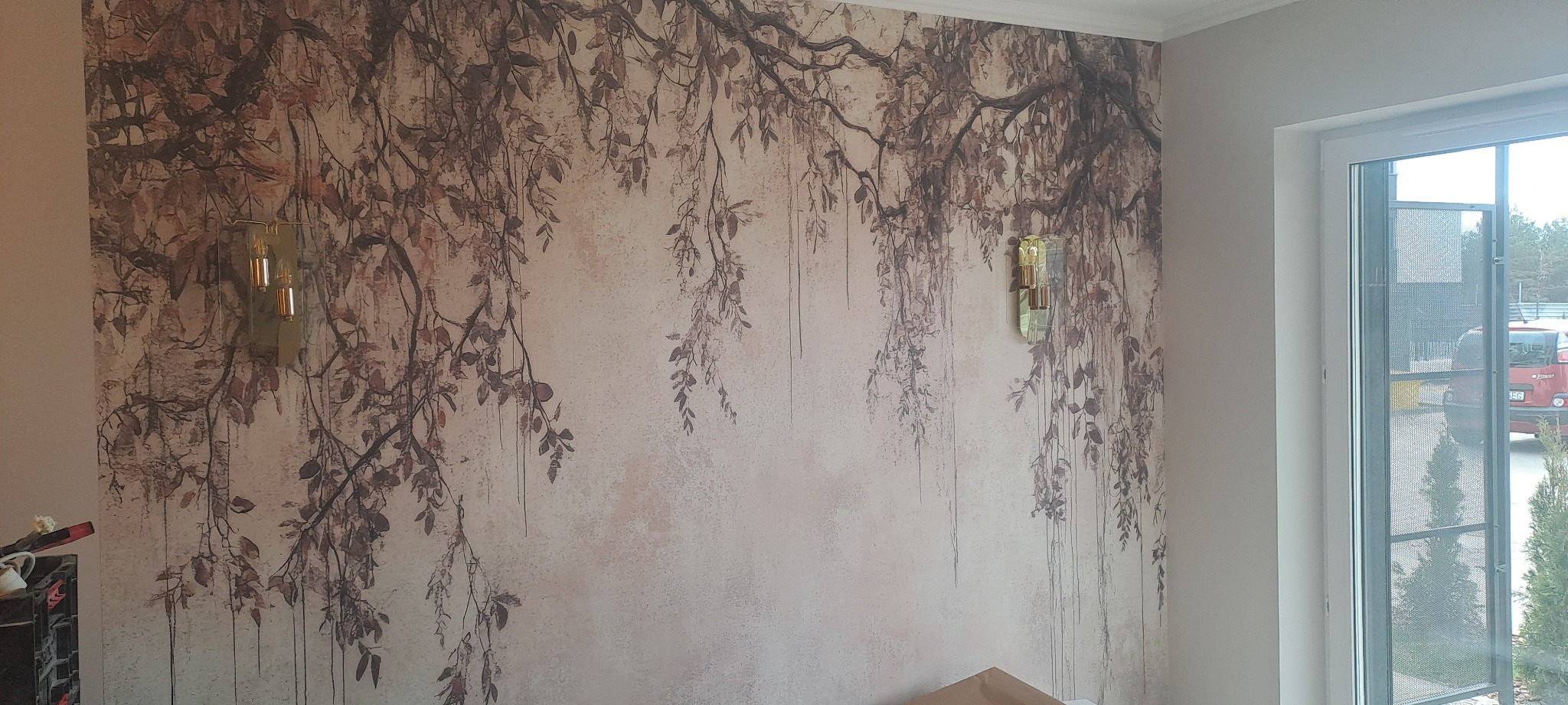 Ściana z tapetą w motywy roślinne, oświetlona dwoma złotymi kinkietami. Widoczne okno z widokiem na zewnątrz. Dekoracyjne wykończenie wnętrza.