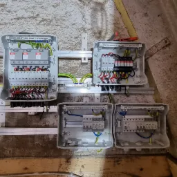 Szare skrzynki elektryczne z okablowaniem i zabezpieczeniami zamontowane na szynie, część instalacji w surowym pomieszczeniu.