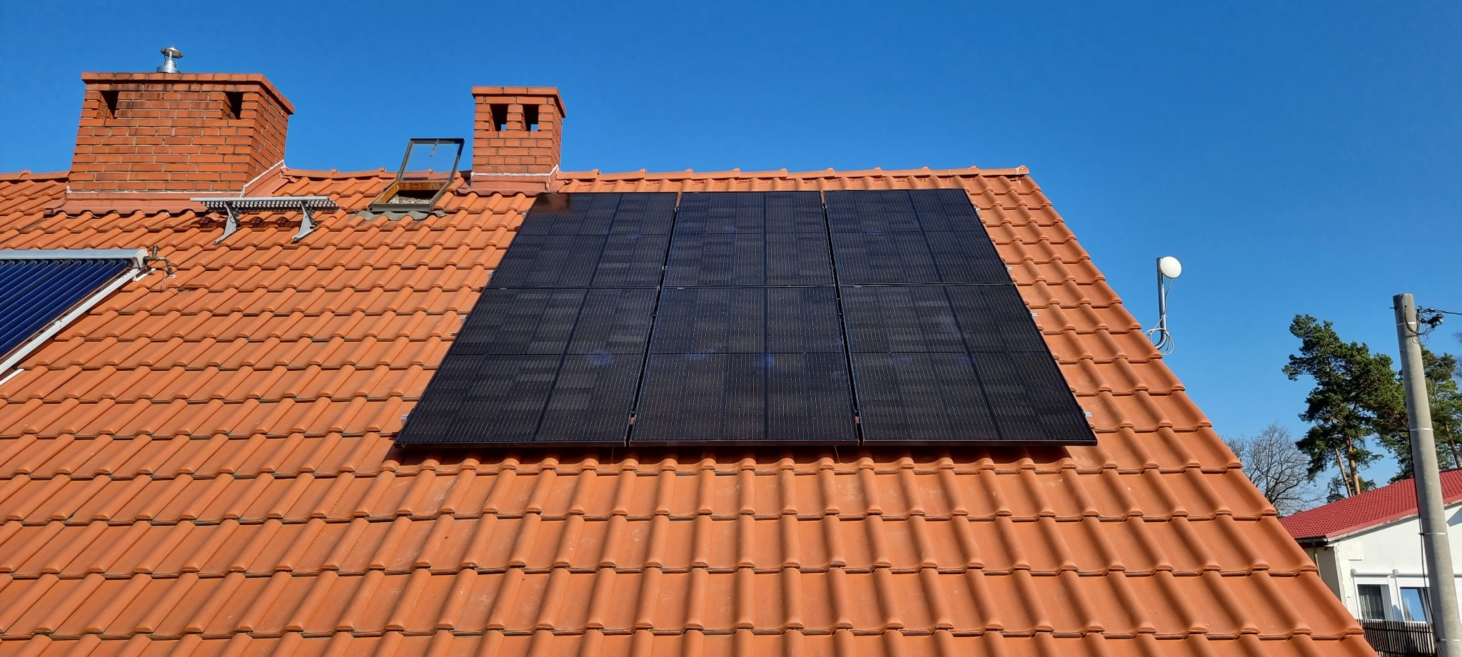 Panele fotowoltaiczne na pomarańczowym dachu domu w Namysłowie, widoczne kominy, antena satelitarna i fragment kolektora słonecznego.