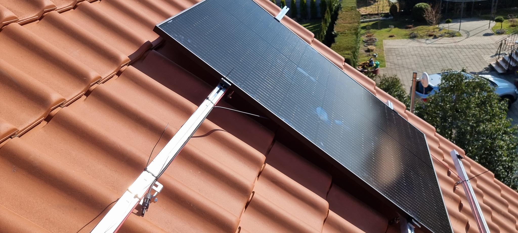 Panel słoneczny zamontowany na dachu z czerwonej dachówki, widoczne elementy montażowe i fragment otoczenia z zielenią i samochodem.