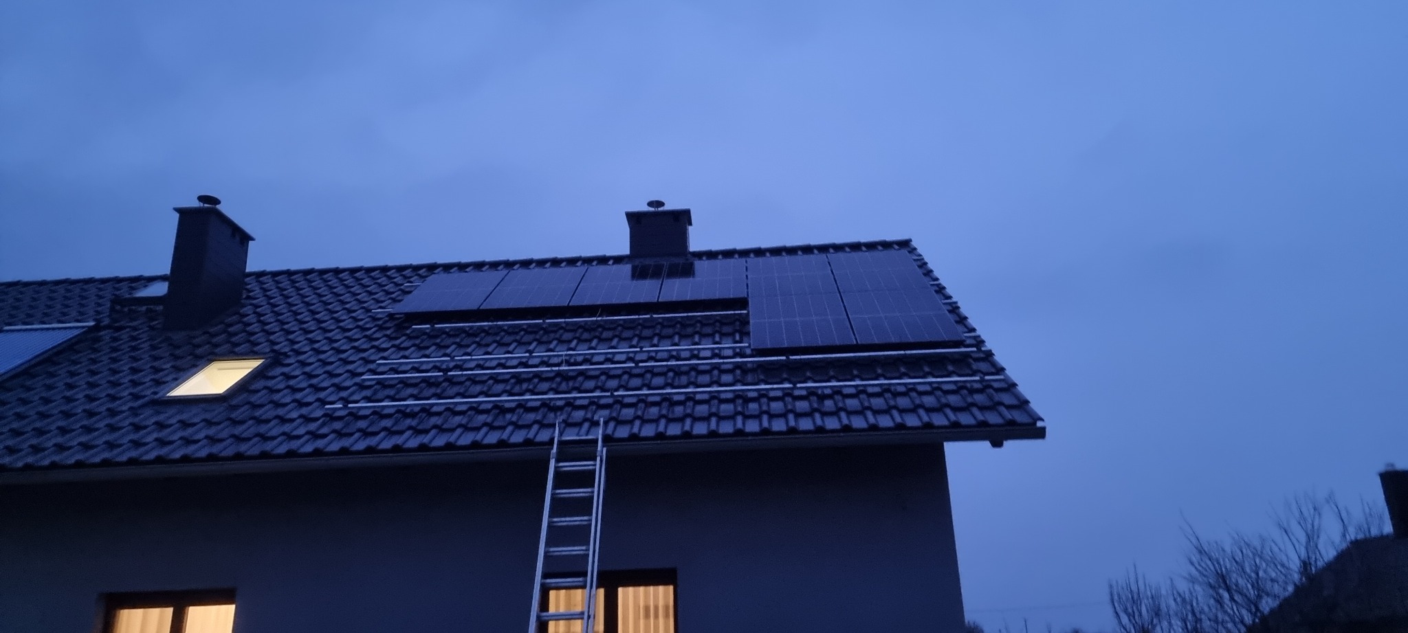 Montaż paneli fotowoltaicznych na dachu domu jednorodzinnego z użyciem drabiny, widoczne szyny montażowe i komin, sfotografowane o zmierzchu.
