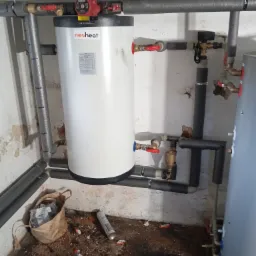 Bojler marki nesheat z podłączonymi rurami i zaworami, widoczny fragment drugiego bojlera, instalacja w piwnicy, na podłodze resztki po montażu.