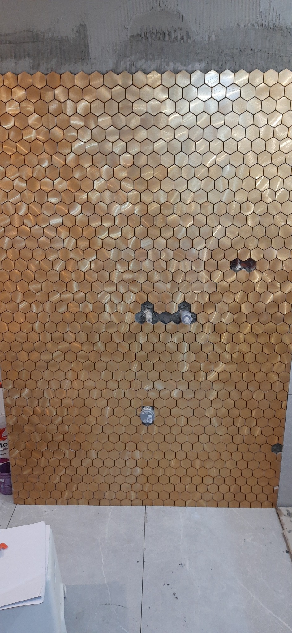 Ściana w trakcie wykańczania z mozaiki heksagonalnej w odcieniach złota, widoczne instalacje wodne i fragment surowego betonu u góry.