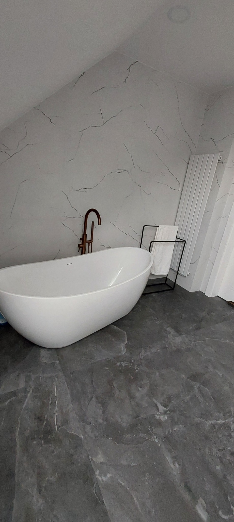 Nowoczesna łazienka na poddaszu z wolnostojącą wanną, baterią w kolorze miedzi i marmurowymi płytkami na ścianach. Grzejnik drabinkowy i ręcznik dodają przytulności.