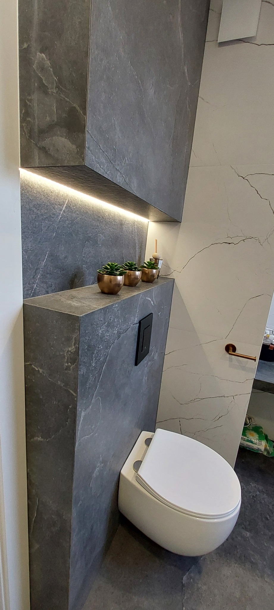 Nowoczesna łazienka z szarą marmurową zabudową, podświetlaną półką z sukulentami i podwieszaną toaletą. Minimalistyczny design z akcentami miedzianymi.