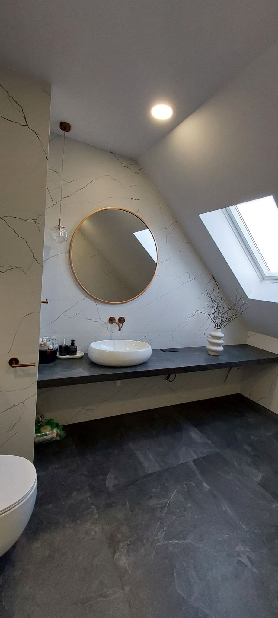 Nowoczesna łazienka na poddaszu z marmurowymi płytkami, okrągłym lustrem w miedzianej ramie i białą umywalką nablatową. Minimalistyczny design z akcentami miedzi.