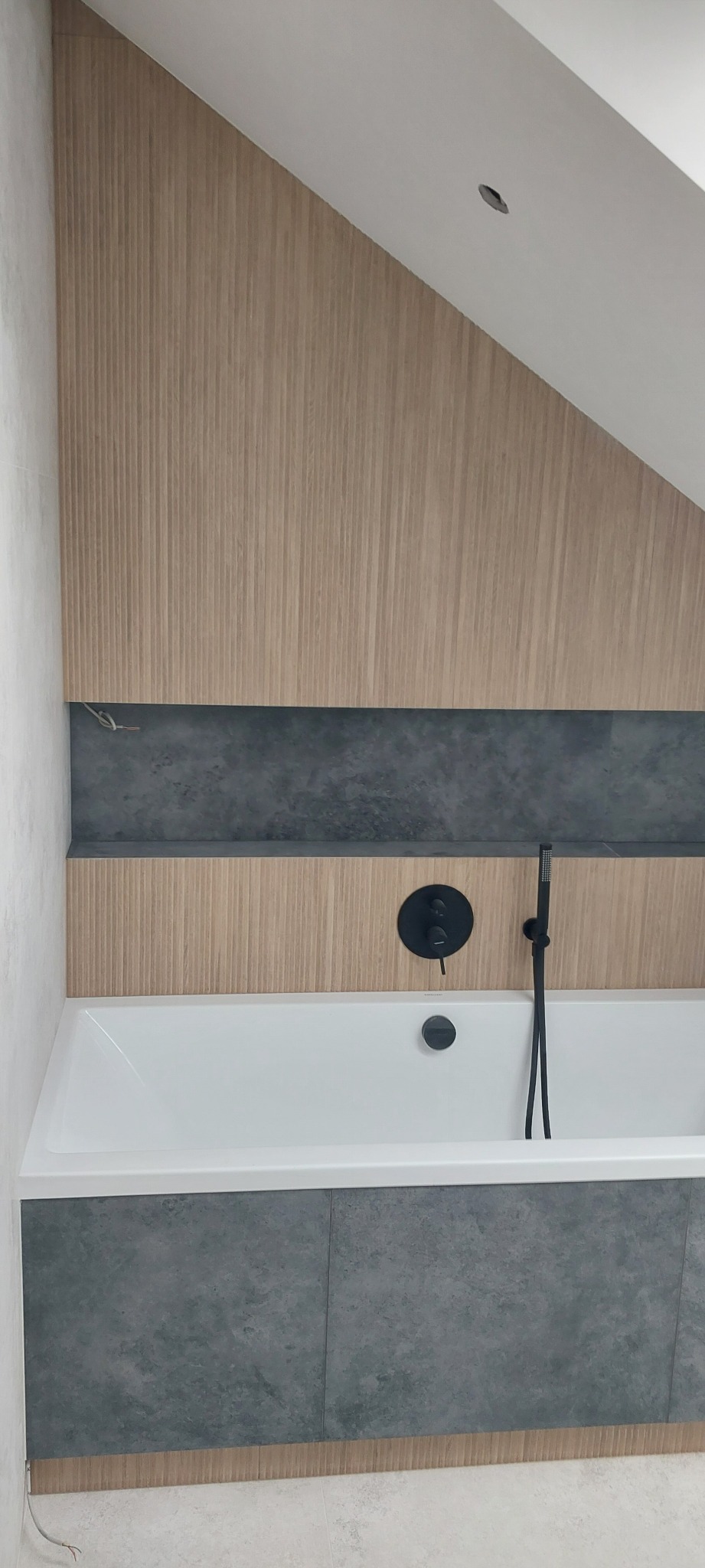 Nowoczesna łazienka na poddaszu: drewnopodobne panele ścienne, szara mozaika nad wanną, czarna armatura. Minimalistyczny design w jasnej tonacji.