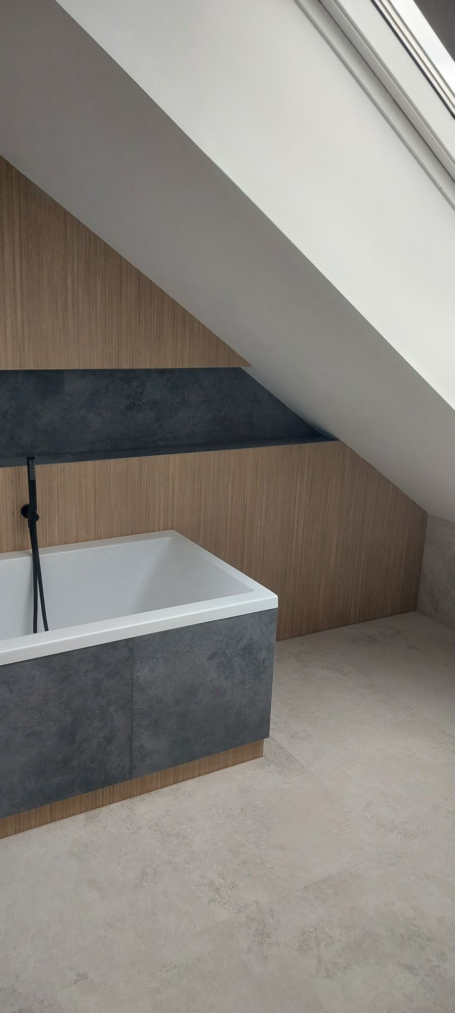 Nowoczesna łazienka na poddaszu z wanną obudowaną szarą płytką, drewnianą boazerią i oknem dachowym. Minimalistyczny design, jasne kolory, kabina prysznicowa z czarną armaturą.
