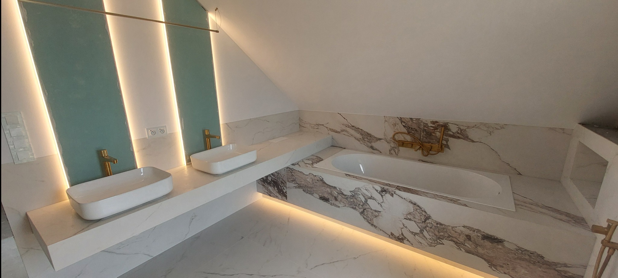 Nowoczesna łazienka na poddaszu z marmurowymi płytkami, podświetlanymi panelami w kolorze morskim i złotą armaturą. Dwie białe umywalki nablatowe.