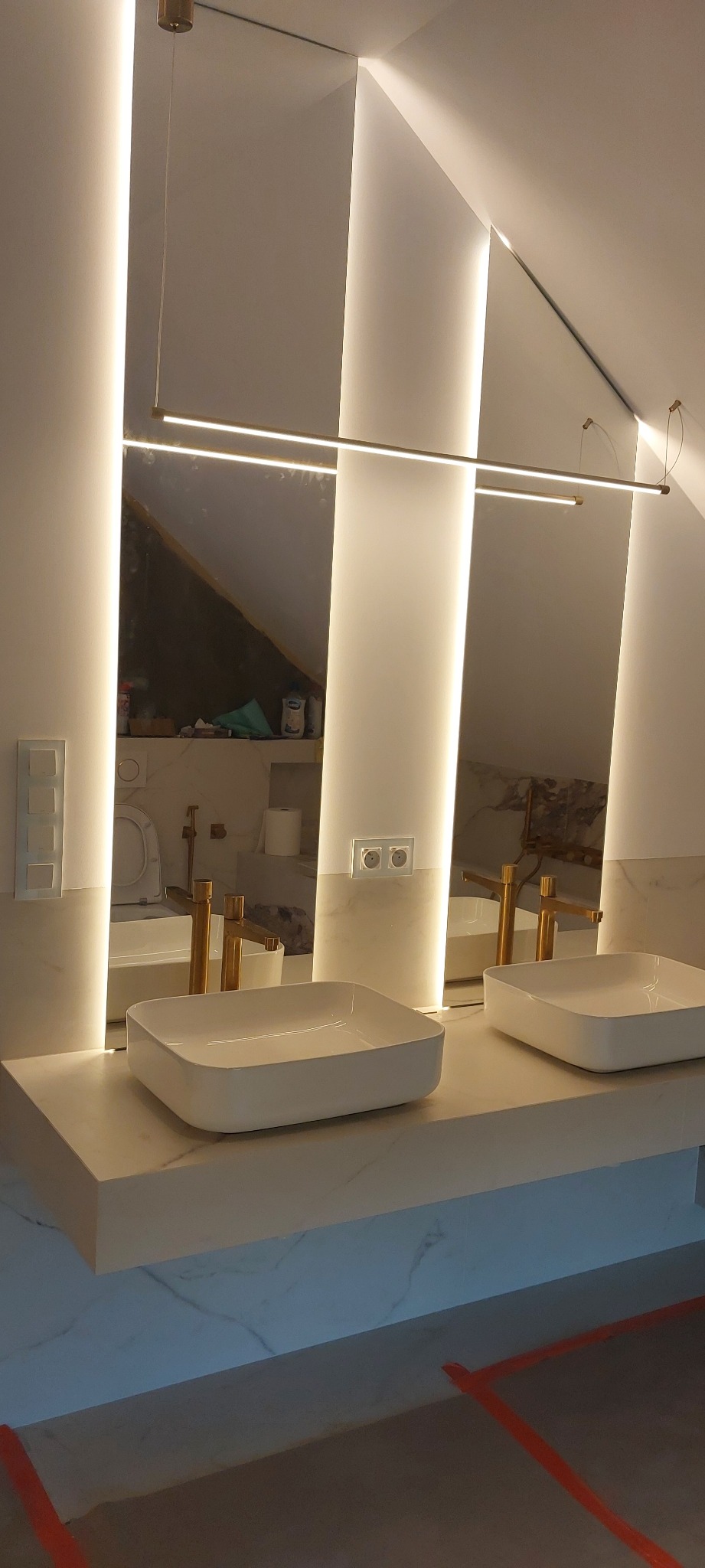 Nowoczesna łazienka w skosach z podświetlanym lustrem i dwiema białymi umywalkami na marmurowym blacie. Widoczne złote baterie i fragment podłogi z zabezpieczeniem.