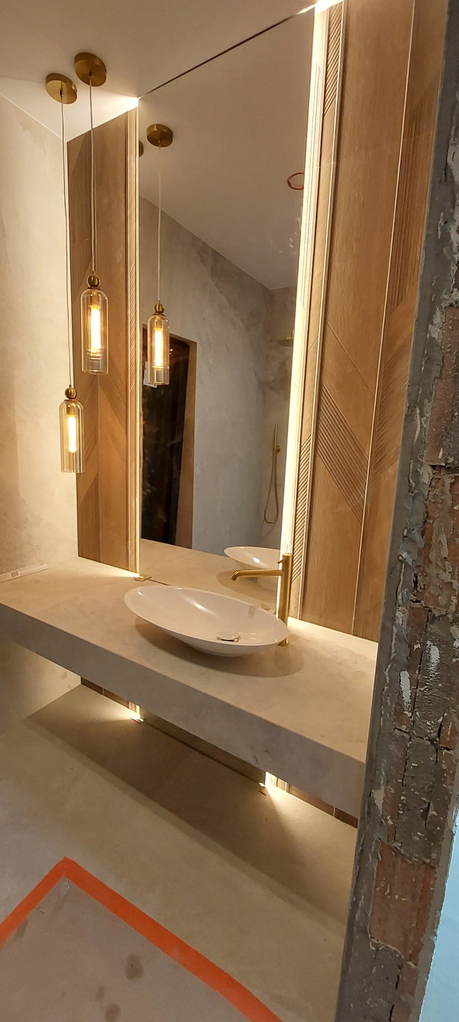 Nowoczesna łazienka w trakcie wykończenia: owalna umywalka, złote lampy wiszące i lustro z drewnianą ramą. Widoczne niedokończone ściany.