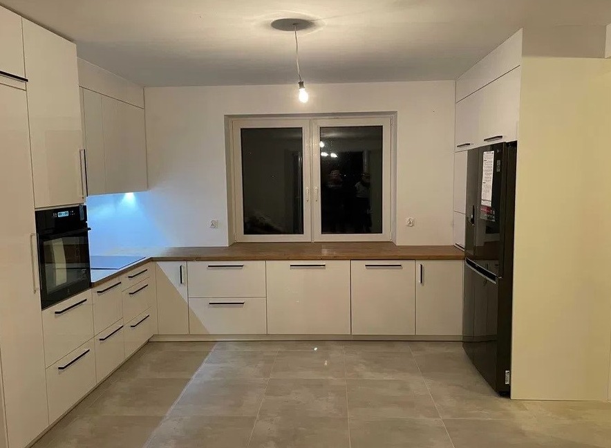 Nowoczesna, biała kuchnia z drewnianym blatem pod oknem, czarnymi uchwytami i czarną lodówką, oświetlona pojedynczą żarówką.