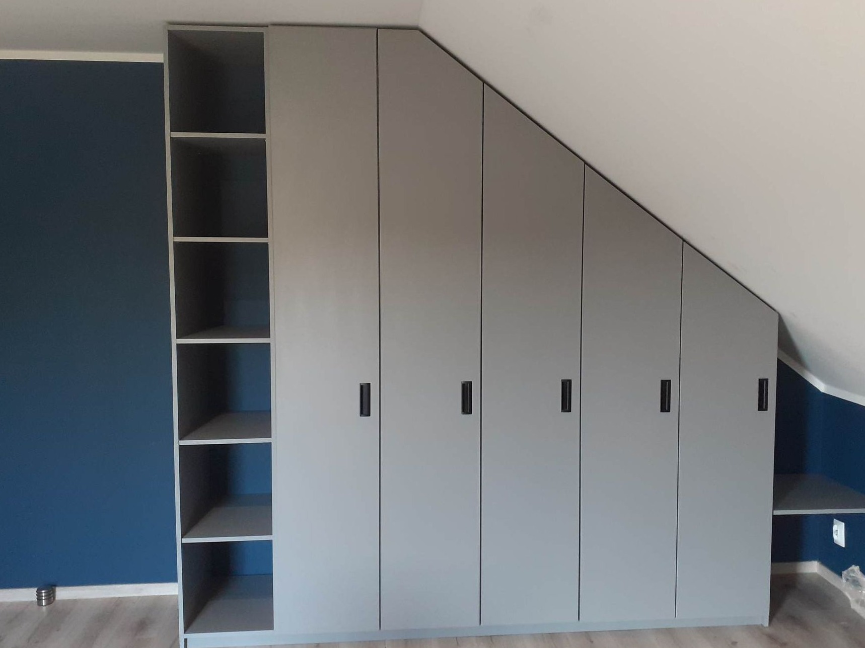 Szara szafa wnękowa na poddaszu ze skośnym sufitem i otwartymi półkami z boku, minimalistyczne czarne uchwyty.